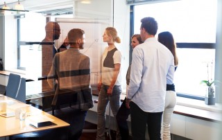 Group of executives standing in conference room gathered around white board