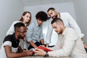 A diverse team of five colleagues gathered around a table reviewing a document together in a bright modern office